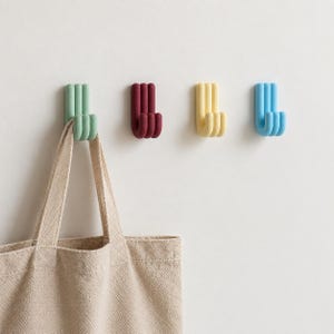 Op de afbeelding: Vier wandhaken in lichtgroen, donkerrood, geel en lichtblauw, elk met een ontwerp met drie tanden. Een beige canvas tas hangt aan de lichtgroene haak, wat de functionele toepassing van deze wandorganizers laat zien.