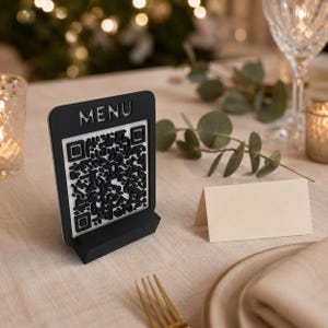 Può includere: Un cartello rettangolare bianco e nero con la parola MENU nella parte superiore e un motivo a codice QR in rilievo al centro. Il cartello è montato su una solida base nera, progettata per stare in piedi su una superficie piana.