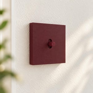 May include: A square, deep burgundy light switch with a matte, textured finish. The switch features a central, cylindrical toggle lever that protrudes from a vertical slot in the middle of the square faceplate, mounted flush against a white wall.
