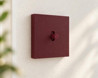 Puede incluir: Un interruptor de luz cuadrado de color burdeos intenso con un acabado mate y texturizado. El interruptor cuenta con una palanca cilíndrica central que sobresale de una ranura vertical en el medio de la placa frontal cuadrada sobre una pared.