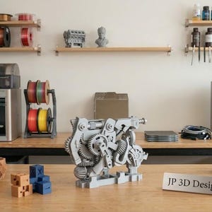 Op de afbeelding: Een 3D-printwerkruimte met twee 3D-printers, filamentrollen op planken en een grijs mechanisch tandwielmodel. Op de tafel staan kleine geometrische plantenbakken, blauwe en beige blokken en een bordje met de tekst JP 3D Design.