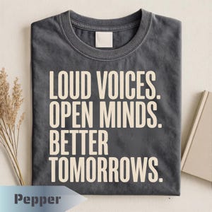 May include: A charcoal gray crew neck t-shirt featuring the cream-colored text LOUD VOICES. OPEN MINDS. BETTER TOMORROWS. printed in a bold, sans-serif font on the front. The shirt has a classic short-sleeve design and a standard ribbed neckline.