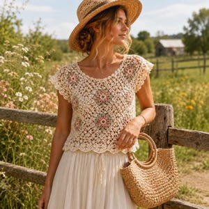 May include: A cream-colored crochet top featuring a repeating floral motif in shades of dusty pink and sage green, paired with a straw hat and a woven straw handbag. The top has short sleeves and a scalloped hem, worn with a long white skirt.
