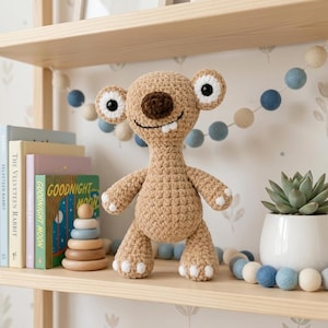Puede incluir: Una criatura de ganchillo de color marrón claro con grandes ojos redondos, una nariz marrón oscura y un solo diente blanco. La figura de peluche tiene una superficie de hilo texturizado, orejas redondeadas y detalles blancos en manos y pies.