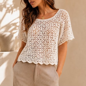 May include: A cream-colored short-sleeve top featuring an intricate openwork crochet pattern with a scalloped hemline and a wide round neckline. The garment is paired with light tan trousers, showcasing a textured, lightweight knit construction.