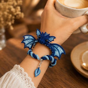 Op de afbeelding: Een armband met een gehaakte donkerblauwe draak met lichtblauwe vleugels en zwarte ogen. De band is gemaakt van gedraaid donker- en lichtblauw koord, afgewerkt met een zilverkleurige metalen ketting en een kleine, bijpassende gehaakte bladbedel.