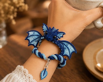 Könnte beinhalten: Ein Armband mit einem gehäkelten dunkelblauen Drachen mit hellblauen Flügeln und schwarzen Augen. Das Band besteht aus einer gedrehten dunkel- und hellblauen Kordel, ergänzt durch eine silberfarbene Metallkette und einen kleinen, passenden Blattanhänger.