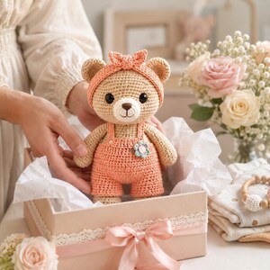 May include: A crocheted teddy bear with light brown yarn, wearing a matching peach-colored headband with a bow and peach-colored overalls. The bear features a small, multi-colored crocheted flower accent on the front of the overalls and black eyes.