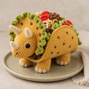 May include: A crocheted triceratops dinosaur toy shaped like a taco. The dinosaur is yellow with three cream-coloured horns and a green frill. The taco shell is yellow with brown spots, filled with brown ground meat, red tomatoes, and green lettuce.