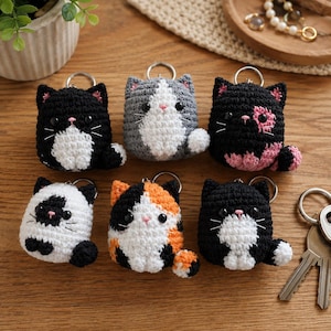 May include: Six crocheted cat keychains featuring various color patterns including black and white, grey and white, black and pink, white and black, calico, and black and white. Each plush cat has black bead eyes, a pink nose, and a metal ring.