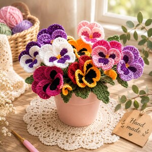 May include: A handmade bouquet of colourful crochet pansies in a pink pot. The flowers feature shades of purple, pink, white, orange, and red with green leaves. The pot sits on a cream doily, with a tag that says "Hand made".