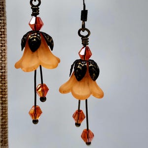 May include: A pair of handcrafted earrings featuring peach-colored flower-shaped pendants with black accents. Each earring has dangling orange crystal beads and dark metal wire detailing. The earrings are displayed on a neutral-toned display stand.