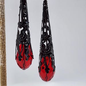 May include: A pair of black filigree earrings with a teardrop shape. The earrings have a detailed, openwork design and feature a faceted, red crystal at the bottom. The earrings are hanging on a neutral-colored display stand.