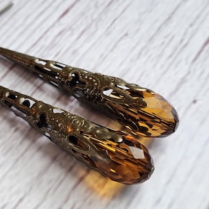 May include: A pair of long dangle earrings featuring faceted amber-colored glass teardrop beads encased in intricate, dark bronze-toned metal filigree cages. The earrings are attached to simple dark metal ear wires for pierced ears.