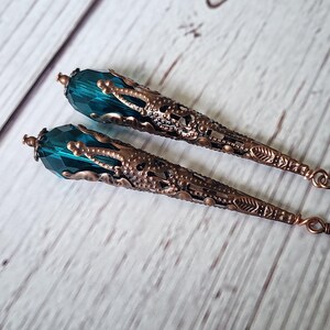 K&ouml;nnte beinhalten: Ein Paar lange, verzierte Ohrringe. Jeder Ohrring hat einen facettierten teal-farbenen Kristall an der Spitze, der mit einem detaillierten, bronzefarbenen Filigran-Kegel verbunden ist. Die Ohrringe haben einen Hakenverschluss.