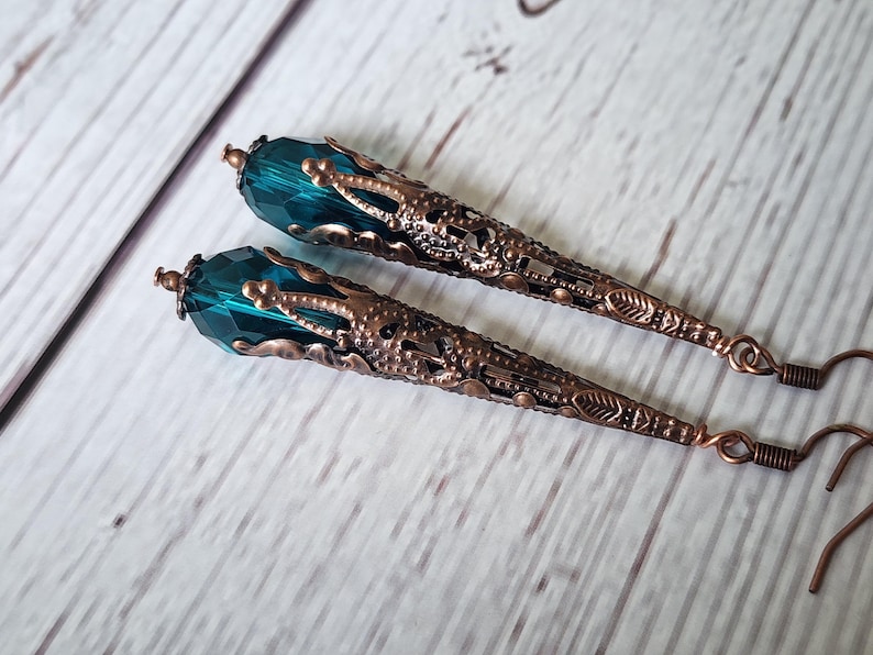 K&ouml;nnte beinhalten: Ein Paar lange, verzierte Ohrringe. Jeder Ohrring hat einen facettierten teal-farbenen Kristall an der Spitze, der mit einem detaillierten, bronzefarbenen Filigran-Kegel verbunden ist. Die Ohrringe haben einen Hakenverschluss.