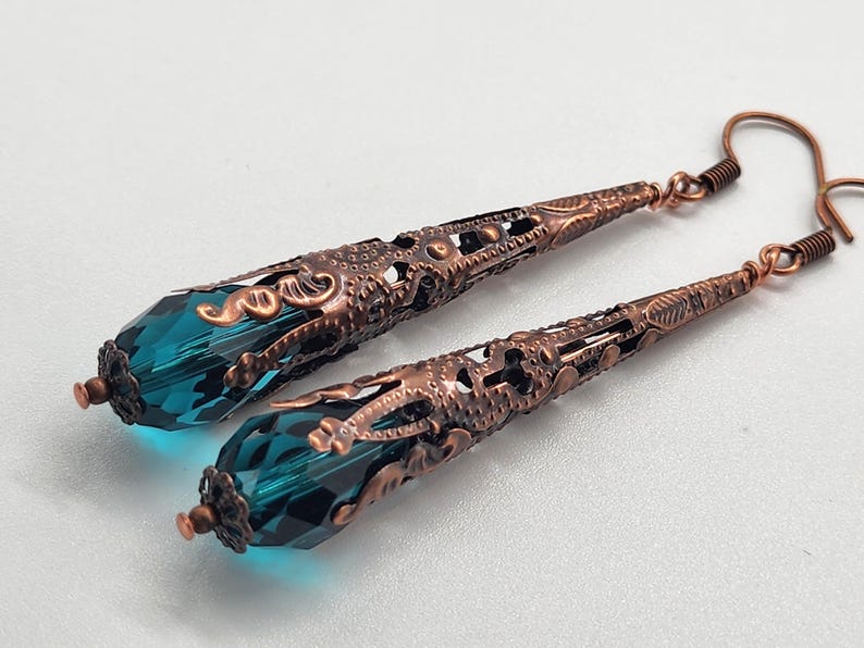 K&ouml;nnte beinhalten: Ohrringe mit tropfenf&ouml;rmigen, teal-farbenen Kristallen. Die Ohrringe haben filigrane Metallarbeiten in Kupferfarbe und dekorative Hakenverschl&uuml;sse. Das Design ist elegant und eignet sich f&uuml;r besondere Anl&auml;sse. Die Ohrringe sind ca. 6 cm lang.