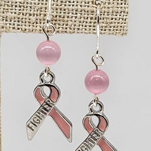 May include: Pink ribbon earrings with the word "Fighter" on them. The earrings are made with pink beads and silver ribbon charms.