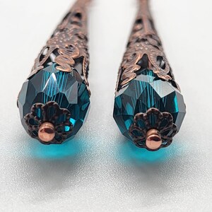 K&ouml;nnte beinhalten: Nahaufnahme von zwei kupferfarbenen Filigranohrringen mit facettierten, teal-farbenen Kristall-Tropfenanh&auml;ngern. Die Ohrringe haben ein detailliertes, offenes Design und eine kleine, kupferfarbene bl&uuml;tenf&ouml;rmige Kappe an der Basis des Kristalls.