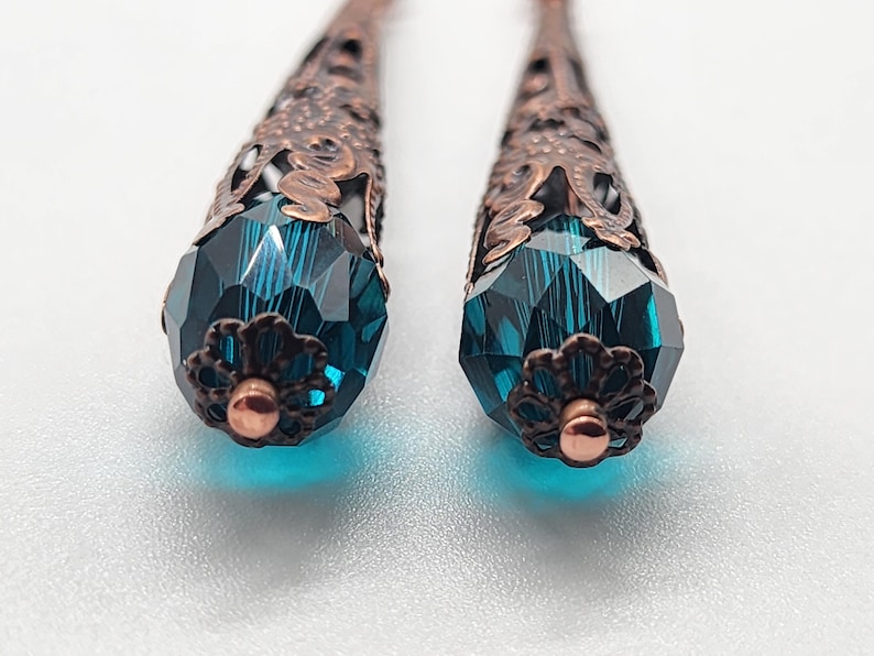 K&ouml;nnte beinhalten: Nahaufnahme von zwei kupferfarbenen Filigranohrringen mit facettierten, teal-farbenen Kristall-Tropfenanh&auml;ngern. Die Ohrringe haben ein detailliertes, offenes Design und eine kleine, kupferfarbene bl&uuml;tenf&ouml;rmige Kappe an der Basis des Kristalls.