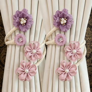 Pode incluir: Laços de cortina florais em tons de roxo e rosa. Os laços apresentam flores de tecido com detalhes de pérolas e são presos a uma corda creme. As cortinas são brancas e o fundo é um tecido estampado.