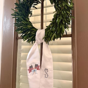Könnte beinhalten: Ein grüner Blätterkranz hängt an einem Fenster und ist mit einem weißen Band verziert. Das Band zeigt gestickte amerikanische Flaggen und ein monogrammiertes "S". Der Kranz hat eine kreisförmige Form und besteht aus grünen Blättern.