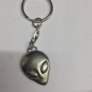 May include: A silver-toned metal keychain featuring a stylized alien head pendant. The pendant is attached to a standard circular split ring by a short metal chain. The alien head has a smooth, rounded surface with two large, almond-shaped eyes.