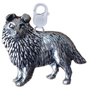 May include: A silver-coloured charm depicting a Collie dog. The charm features detailed fur texture and a lobster clasp for attachment. The dog is in a standing pose, with its head turned. The charm is likely made of metal.