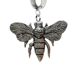 Könnte beinhalten: Ein silberfarbener Bienenanhänger mit detaillierten Flügeln und Körper. Der Anhänger hat oben eine Schlaufe zum Befestigen an einem Armband oder einer Halskette. Die Biene ist ein beliebtes Symbol in der Schmuckherstellung.