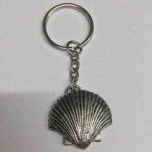May include: A silver-toned metal keychain featuring a textured scallop shell charm attached to a circular split ring by a short chain. The shell has distinct vertical ridges and a fan-like shape, with a metallic finish that reflects light.