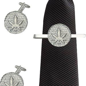 Puede incluir: Una corbata negra con textura acanalada diagonal, acompañada de un pasador de corbata plateado y dos gemelos a juego. Cada pieza presenta un diseño circular con fondo texturizado y un emblema de hoja de cannabis en relieve en el centro.