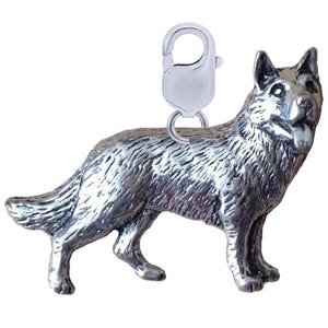 May include: A silver German Shepherd dog charm with a lobster clasp. The dog is shown standing with its tongue visible. The charm has a textured surface, giving it a detailed appearance.