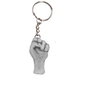 Pode incluir: Um porta-chaves com uma mão esculpida em cinzento claro numa posição de punho fechado. A mão está presa a um anel de metal prateado por uma curta corrente de elos metálicos. O design capta os detalhes anatómicos de um punho fechado.