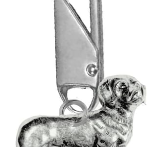 Puede incluir: Un llavero plateado con un dije detallado de perro salchicha. El dije está unido a un cierre de metal, diseñado para asegurar llaves u otros objetos pequeños. El dije de perro es una escultura en miniatura de un perro salchicha.