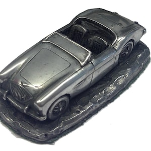 May include: A silver miniature classic convertible car, with detailed features including a front grill, headlights, and interior seats. The car is mounted on a textured, dark grey base, creating a vintage aesthetic.