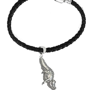 Könnte beinhalten: Ein schwarzes geflochtenes Lederarmband mit einem silberfarbenen Alligator-Anhänger. Das Armband hat einen silbernen Verschluss und einen dekorativen Anhänger. Der Alligator-Anhänger ist detailliert und verleiht dem Schmuck eine einzigartige Note.