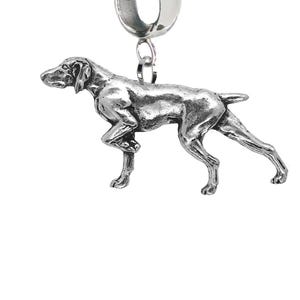 Könnte beinhalten: Ein silberfarbener Metallanhänger in Form eines stehenden Hundes in Seitenansicht. Der Anhänger hat eine glatte, reflektierende Oberfläche und ist an einer großen, runden silberfarbenen Öse befestigt, die zum Auffädeln auf ein Armband oder eine Kette dient.