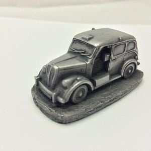 May include: A small, silver-colored vintage taxi cab figurine. The miniature car has detailed features, including headlights, a windshield, and doors. It sits on a textured, rectangular base, and is a collectible item.