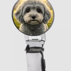 May include: A circular badge clip featuring a detailed illustration of a fluffy grey dog with dark eyes and a black nose. The dog is set against a soft yellow and green outdoor background. The clip is made of polished silver-toned metal.