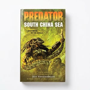 May include: A paperback book titled Predator: South China Sea, Nightmare in the Jungle by Jeff VanderMeer. The cover features a green and yellow jungle scene with a dark, armored creature holding a large snake, with bold orange and white text.