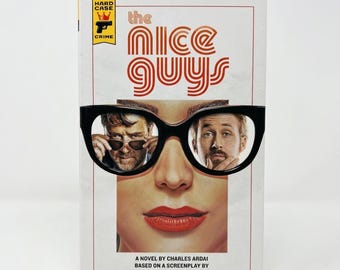 May include: A paperback book titled The Nice Guys by Charles Ardai, based on a screenplay by Shane Black and Anthony Bagarozzi. The cover features a graphic of black eyeglasses framing two men's faces, with a Hard Case Crime logo at the top.