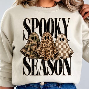 May include: A cream-coloured crewneck sweatshirt featuring the black text SPOOKY SEASON. Between the words are three ghost illustrations: one with a green camouflage pattern, one with a brown leopard print, and one with a tan checkerboard pattern.