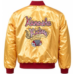 Pode incluir: Uma jaqueta bomber de cetim dourado com gola, punhos e cós canelados em vermelho escuro. A parte de trás apresenta o texto bordado Kenosha Kickers em rosa e branco, acompanhado por uma pauta musical, uma clave de sol e um acordeão.