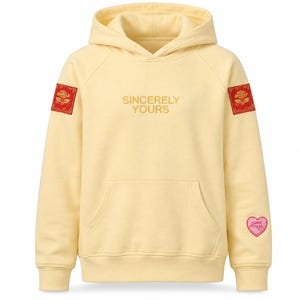 May include: A pale yellow hooded sweatshirt featuring the text SINCERELY YOURS embroidered in the centre. The sleeves have red rectangular patches with gold rose designs, and a pink heart-shaped patch with text is on the lower left sleeve.