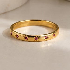 May include: A gold band ring featuring a series of evenly spaced star-shaped engravings, each set with a small, round, vibrant pink gemstone. The metal surface has a polished, reflective finish that catches the light, highlighting the star motifs.