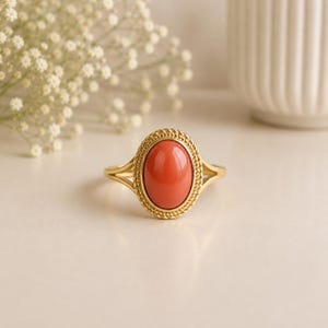 May include: A gold ring featuring a smooth, oval-shaped coral-colored cabochon stone set in the center. The stone is surrounded by a twisted rope-style metal border, and the band splits into two thin, polished gold arms on each side.