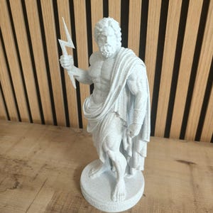 Può includere: Statua grigia chiara di Zeus, il dio greco del cielo e del tuono, che tiene in mano un fulmine. La statua è su una base rotonda e ha un drappo sulla spalla. La statua è alta circa 25 cm.