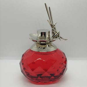 Pode incluir: Um frasco de perfume de vidro vermelho facetado com uma tampa prateada em forma de flor. Uma pequena figura de fada prateada com asas está pousada na borda da tampa. O texto Van Cleef & Arpels está gravado no colar prateado.