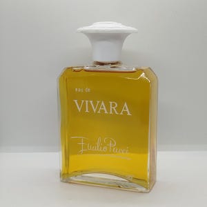 Op de afbeelding: Een rechthoekige glazen parfumfles gevuld met amberkleurige vloeistof, voorzien van een witte dop en de tekst "eau de VIVARA" en "Emilio Pucci" op de voorzijde. De fles heeft een gebogen onderkant en transparante glazen zijkanten.