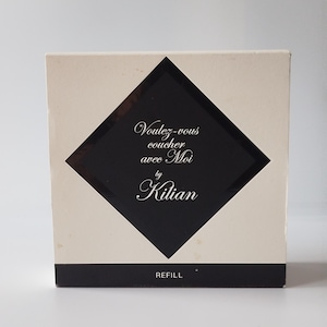 Peut inclure: Une boîte carrée blanc cassé avec un losange noir central. À l'intérieur du losange, le texte indique "Voulez-vous coucher avec Moi by Kilian" en écriture blanche. Une bande horizontale noire en bas contient le mot "REFILL".